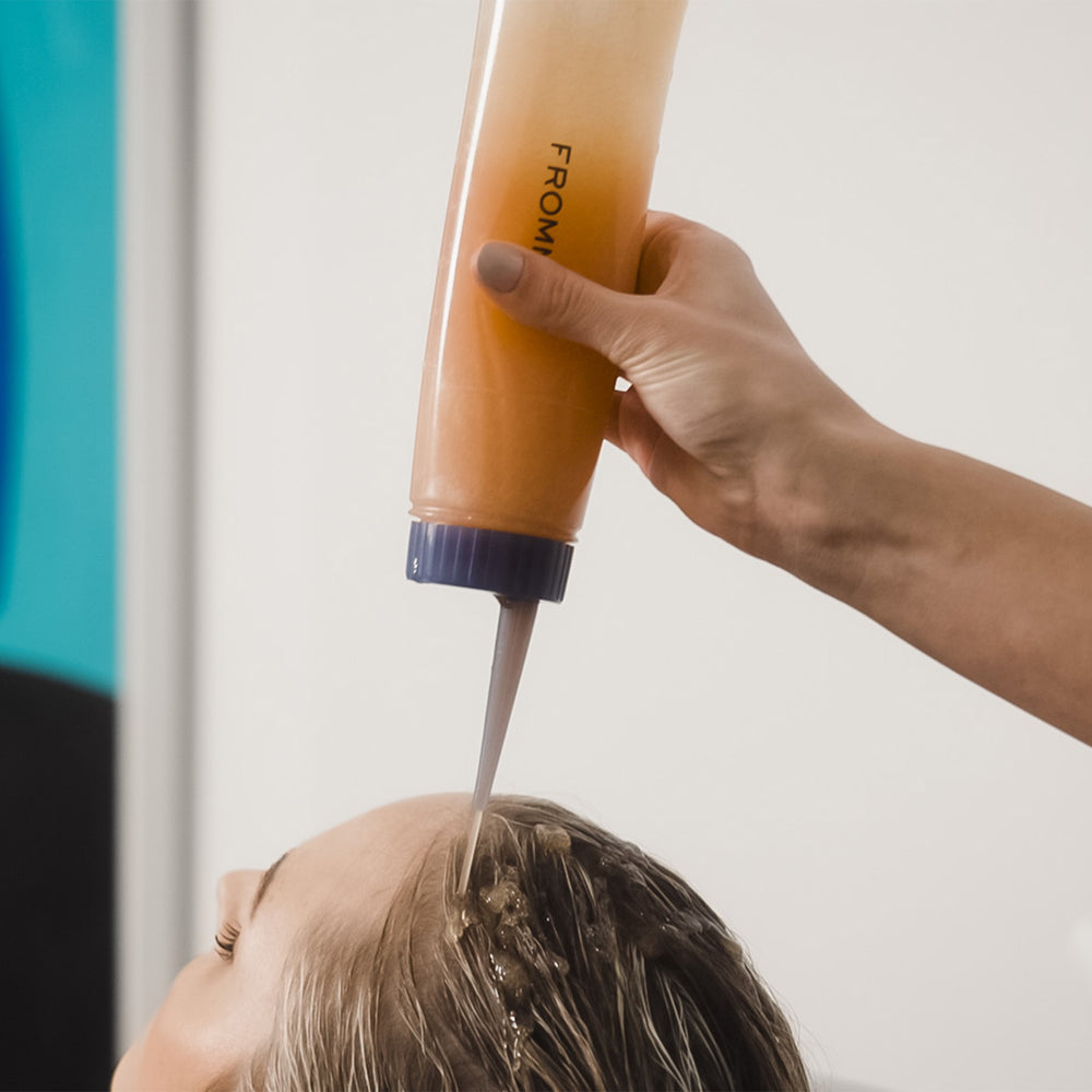 A hand squeezes an application bottle of hair coloring onto a person's scalp. The bottle has a dark blue cap and the brand name 'FROMM' printed vertically on the side.