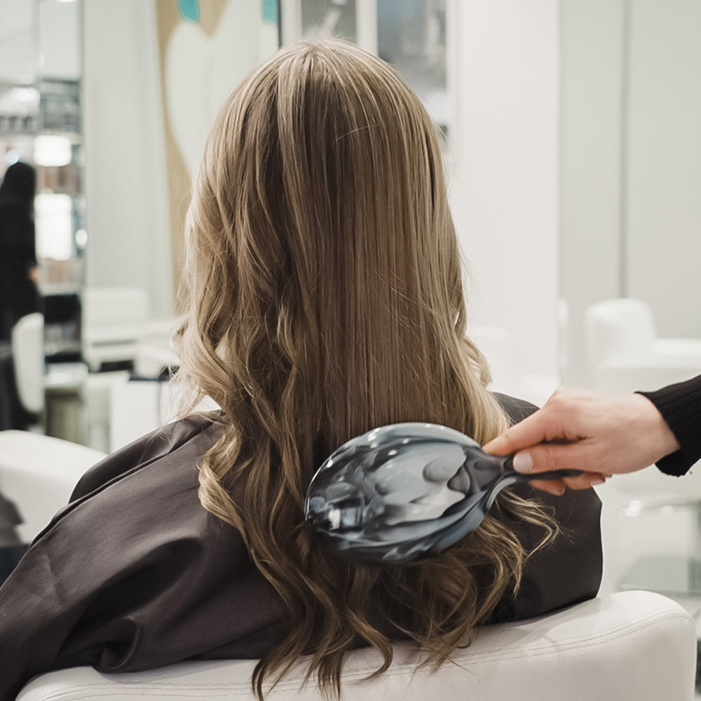 A woman seated in a salon chair seen from behind, with long wavy light brown hair and a hand using a dark paddle brush to comb through the hair in a bright salon interior.