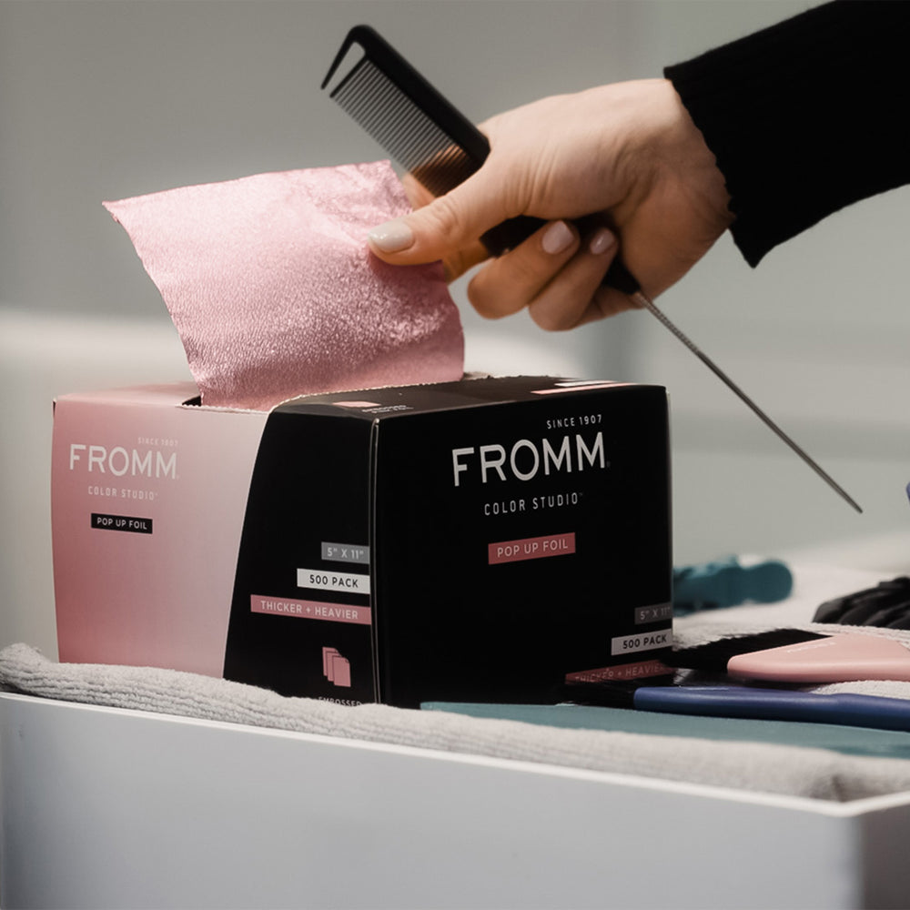 A hand pulls a sheet of pink hair extension from the box of FROMM Pop Up Foil. The box is black and pink, and the hair extension sheet is a shimmering light pink.