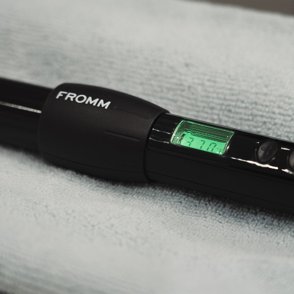 A close-up of a black FROMM hair tool with a digital temperature display showing 370 degrees Fahrenheit, resting on a textured light gray surface.