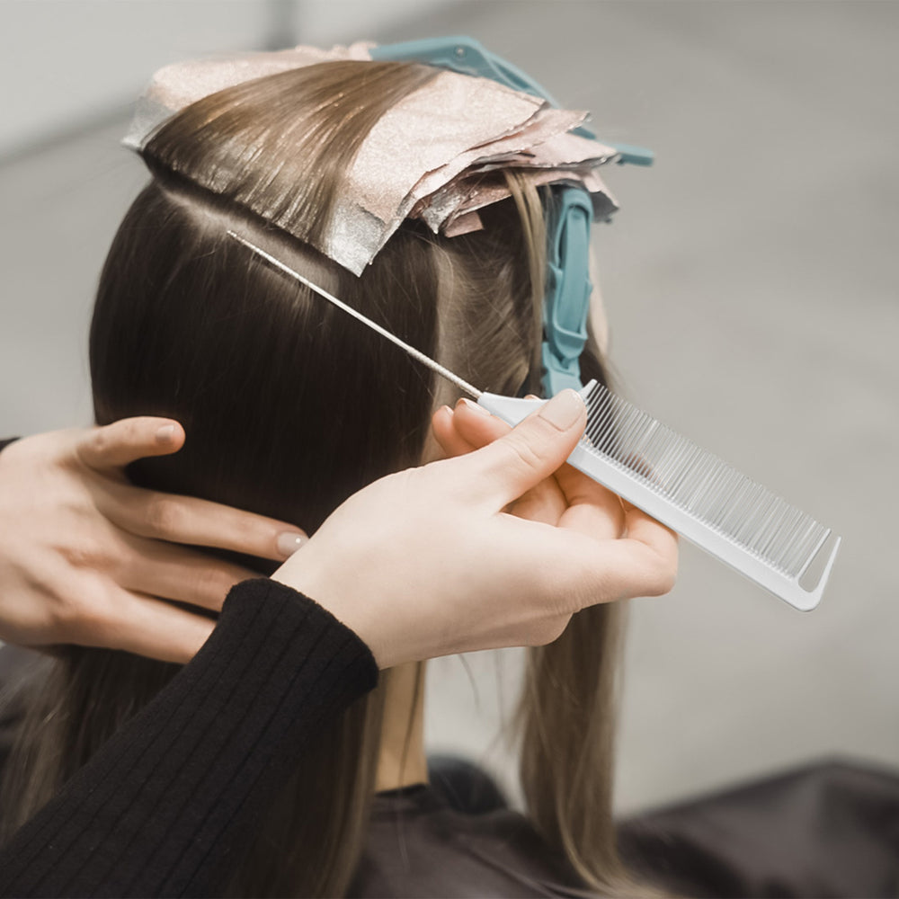 A close-up of a woman's head with hair foils in it during a hair salon session, with a stylist using a white comb to section dark brown hair. 