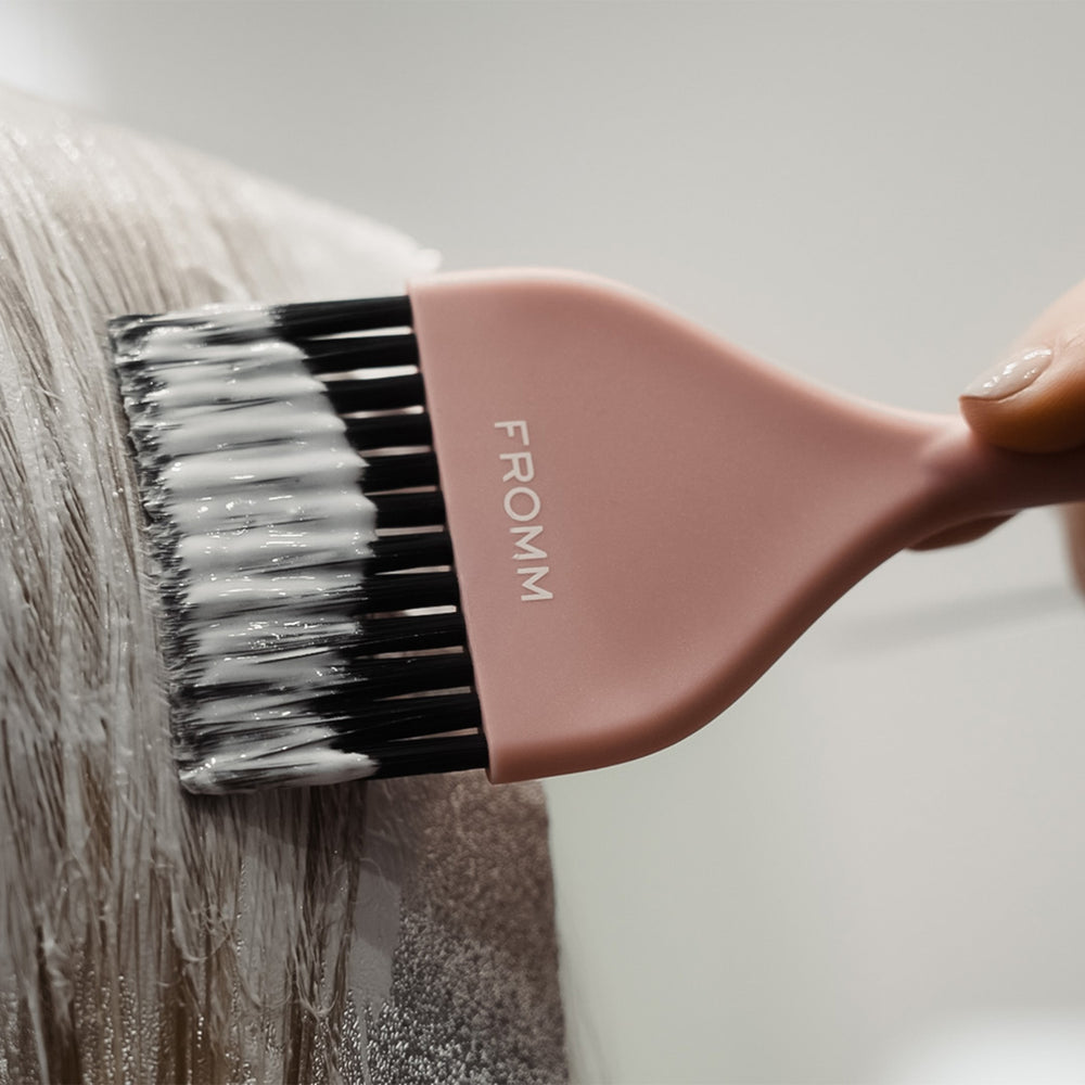 A close-up of a pink hair color brush with black bristles, coated with white hair developer, being used to apply the product to light brown hair. The brand name 'FROOM' is printed on the brush handle.