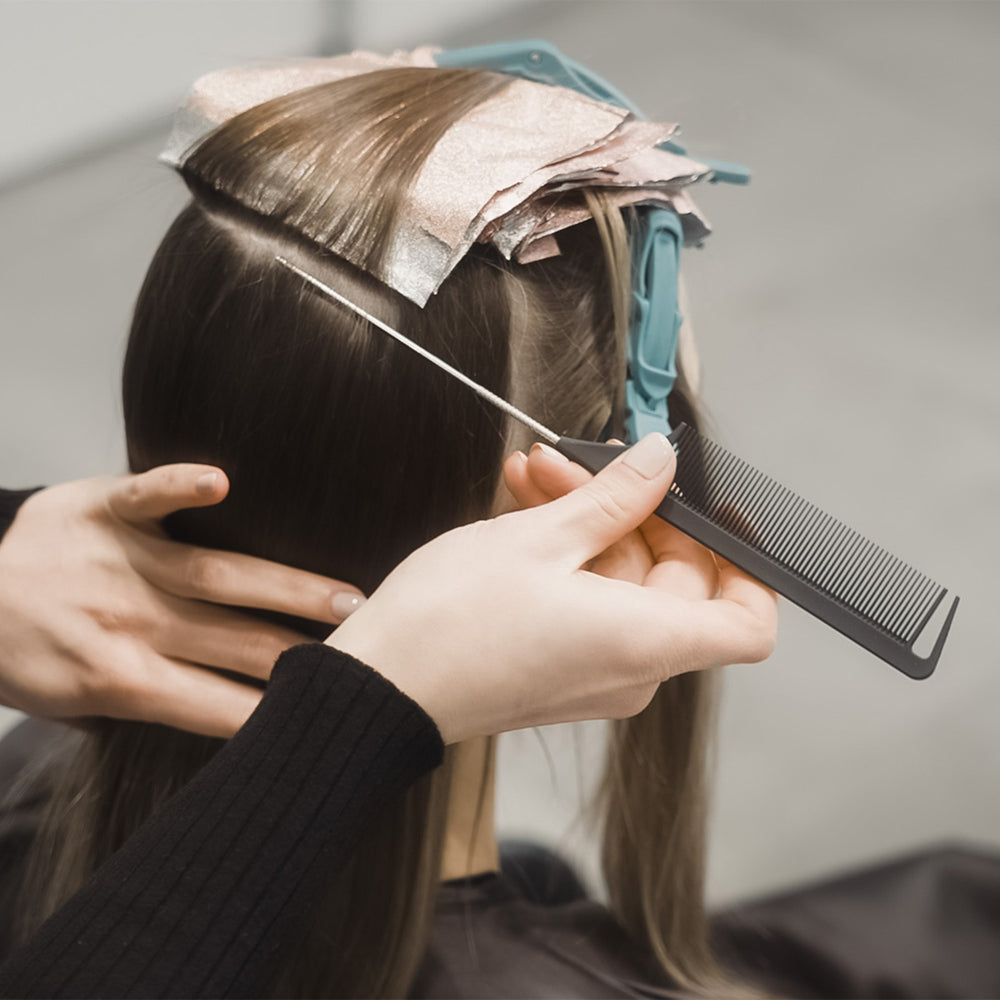 A close-up of a hairstylist using a fine-tooth comb to section a person's dark brown hair, with hair foils applied on top and teal clips holding the hair in place.