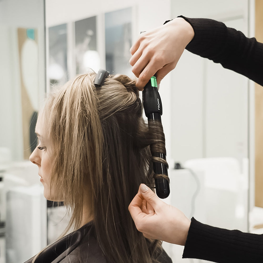 A curling iron with a black barrel and green accent is being used on a client's hair in a salon setting. The tool has the brand name 'FROMM' visible on the side.