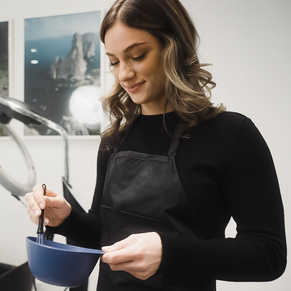 A hair colorist wearing a black long-sleeve top and a black apron, holding a blue color bowl and a whisk in a salon.